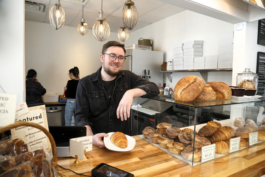 Artistan Bakery owner Sean standing behind a counter with bread, croissants, and more in a bakery